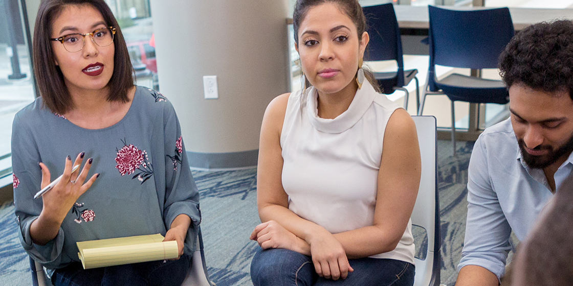 Two mothers sitting next to each other in a meeting. The mother on the left is talking while holding a legal pad in her lap.