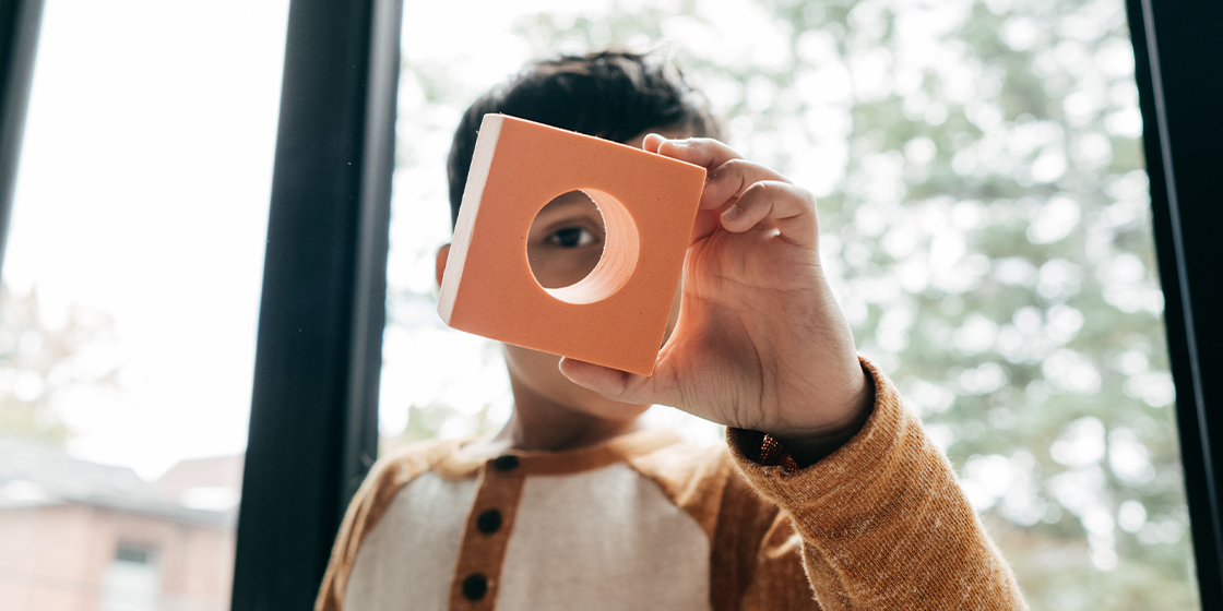 Young child looking at the camera through a square foam block with a circle cut out of the center.