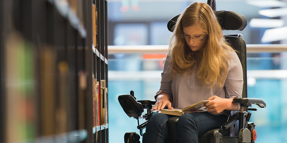 Teenage girl seated in a wheelchair, reading a book in a library.